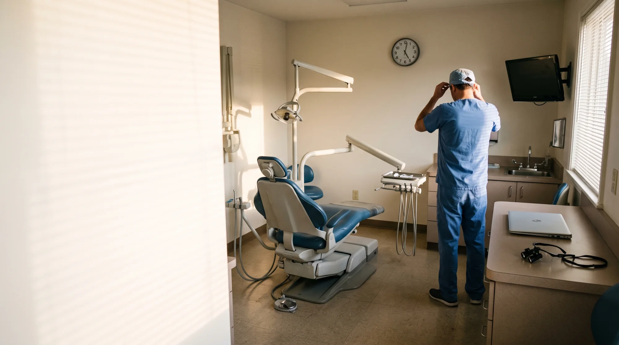 A dentist in scrubs and a cap standing in his own operatory after the last patient, hands behind his head, the dental chair empty and the wall clock past the end of the day.
