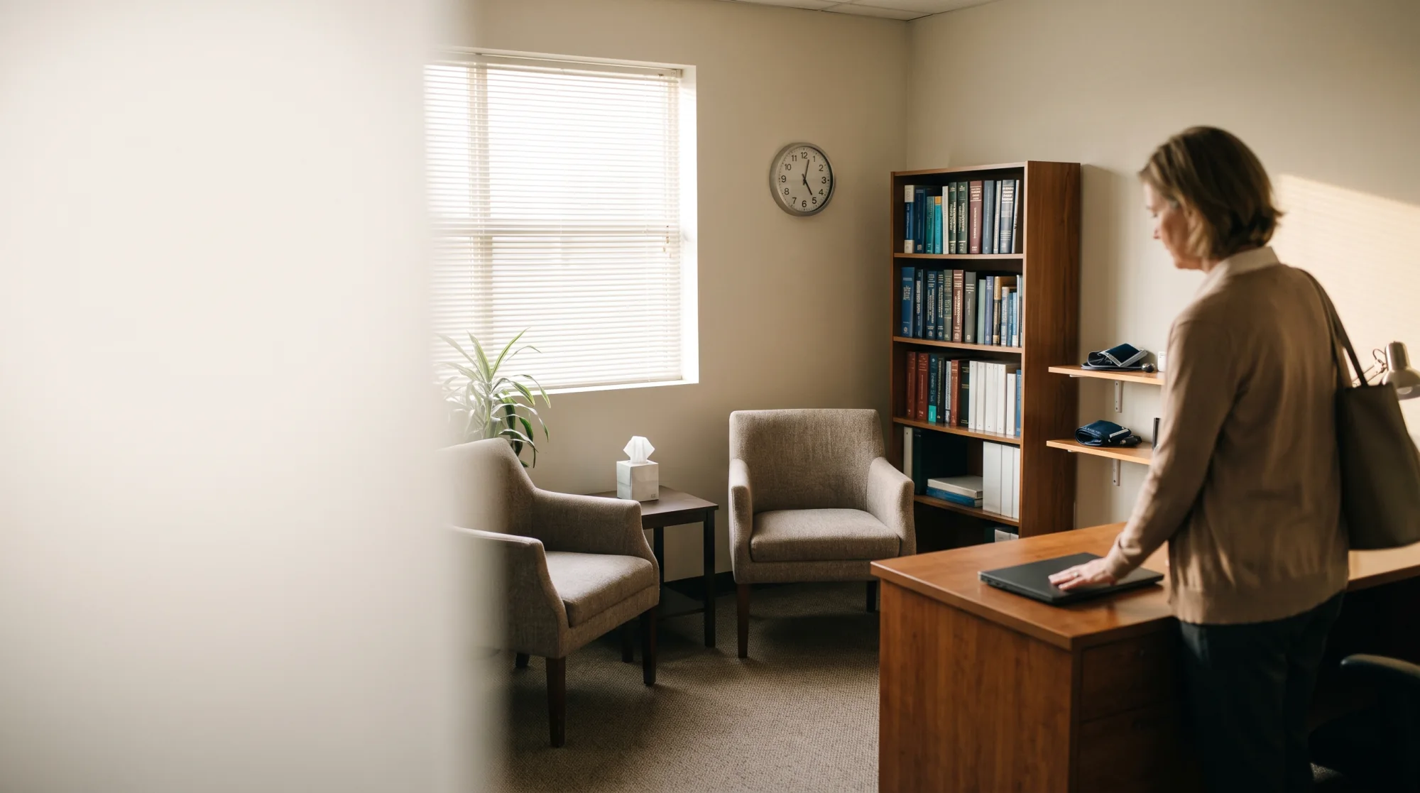 A psychiatrist standing at her desk with one hand on a closed laptop, ready to leave, the two chairs of the consulting room empty and the wall clock past five.